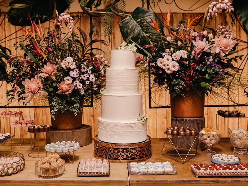 Mesa de dulces de boda con pastel de varios pisos y arreglos florales sobre fondo de madera iluminado
