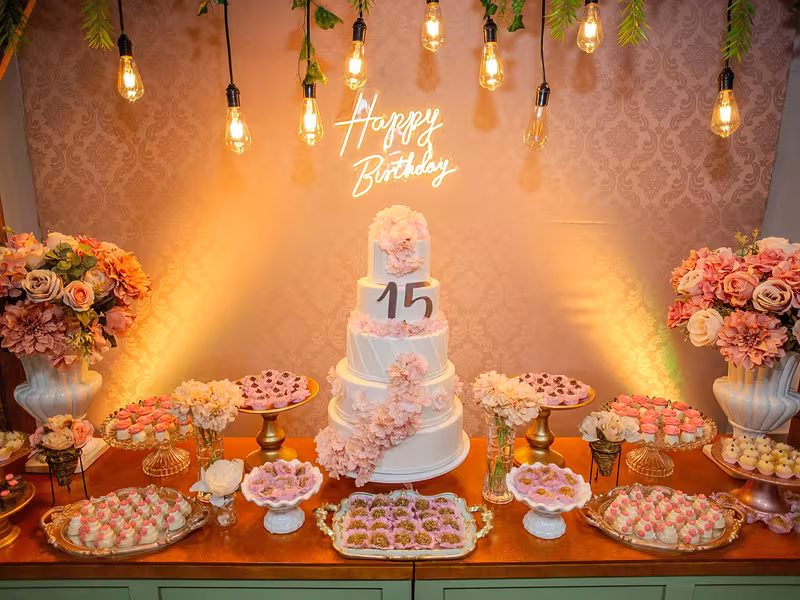 Mesa de dulces de XV años con pastel, focos colgantes y letrero luminoso al fondo